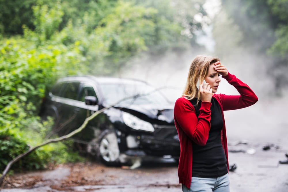 A young woman with smartphone by the damaged car after a car accident, making a phone call.