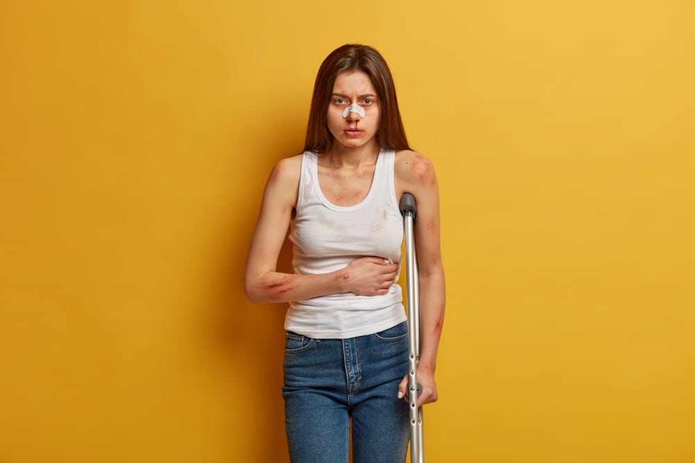 Upset woman has bad feeling after disaster or slip and fall accident, suffers from stomachache, has depressed expression, wears sticky bandage on nose, stands with crutches. Injured cyclist indoor